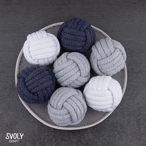 May include: A collection of decorative rope knots in shades of white, gray, and navy blue, arranged on a gray plate. The knots are round and tightly woven, showcasing a nautical or coastal theme. The SVOLY CRAFT logo is visible in the bottom left corner.