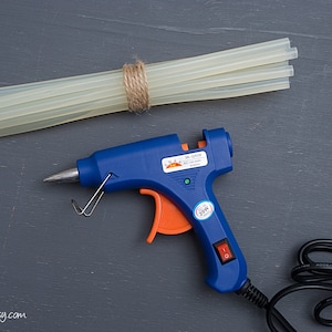 May include: A blue glue gun with an orange trigger and a black power cord. The glue gun is plugged in and ready to use. There are several clear glue sticks bundled together with twine.