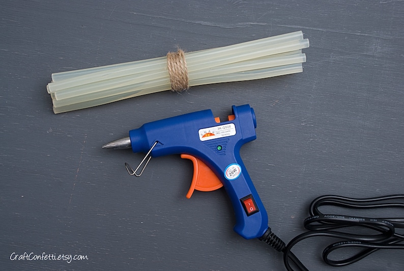 May include: A blue glue gun with an orange trigger and a black power cord. The glue gun is plugged in and ready to use. There are several clear glue sticks bundled together with twine.