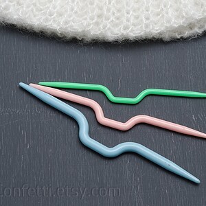 Puede incluir: Tres ganchillos de crochet de pl&aacute;stico en diferentes tama&ntilde;os y colores: azul claro, rosa y verde. Los ganchillos est&aacute;n dispuestos en una fila sobre una superficie gris oscuro.
