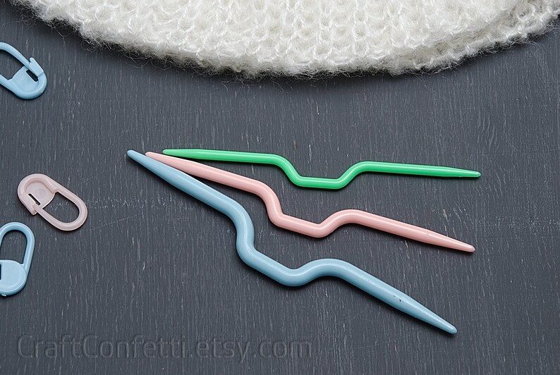 Puede incluir: Tres ganchillos de crochet de pl&aacute;stico en diferentes tama&ntilde;os y colores: azul claro, rosa y verde. Los ganchillos est&aacute;n dispuestos en una fila sobre una superficie gris oscuro.