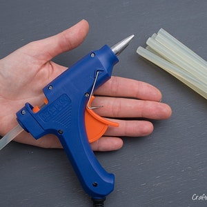May include: A blue and orange glue gun with a clear glue stick. The glue gun is on a person's hand. There are multiple clear glue sticks bundled together with twine.