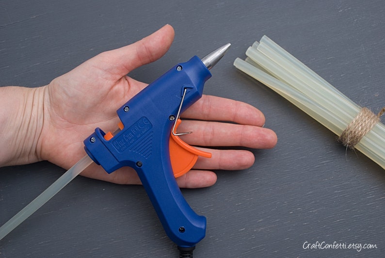 May include: A blue and orange glue gun with a clear glue stick. The glue gun is on a person's hand. There are multiple clear glue sticks bundled together with twine.