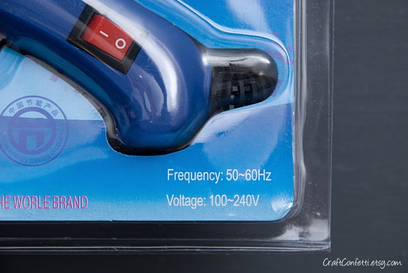 May include: Close-up of a blue and black glue gun with a red on/off switch. The packaging indicates a frequency of 50-60Hz and a voltage of 100-240V.