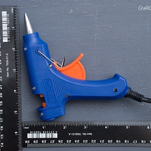 May include: A blue hot glue gun with an orange trigger and a black power cord. The glue gun is measured against a black ruler.