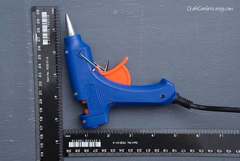 May include: A blue hot glue gun with an orange trigger and a black power cord. The glue gun is measured against a black ruler.
