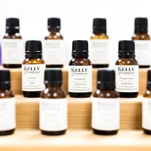 May include: A collection of ten small amber glass bottles with black caps and white labels. The labels have the text "Kelly & Company" and list the essential oil type, origin, and website address. The bottles are arranged on a wooden shelf.
