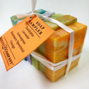 May include: A set of six bars of soap wrapped in white twine and tied with a tag that reads "SOAP SAMPLER" and lists the scents: Eucalyptus, Lavender, Lemongrass, Rosemary Spearmint, Orange. The tag is from Kelly & Company.