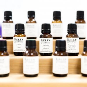 May include: A collection of small amber glass bottles with white labels. The labels have the text "Kelly & Company" and list the essential oil name, origin, and volume. The bottles are arranged on a wooden shelf.