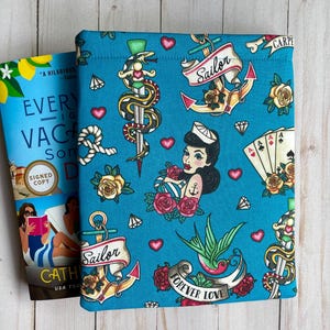 May include: A teal fabric book sleeve featuring a repeating pattern of traditional tattoo-style illustrations including anchors with the word Sailor, roses, hearts, diamonds, playing cards, snakes wrapped around daggers, and swallows with the text Forever Love.