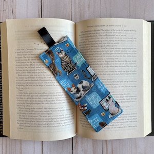 May include: A blue fabric bookmark with a black ribbon. The bookmark features a repeating pattern of cats, coffee cups, and text bubbles with phrases like "Calm Down" and "Degh Kuly".
