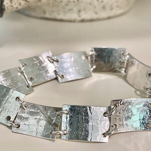 May include: A silver bracelet featuring rectangular, textured metal links connected by small silver rings. The bracelet has a hammered appearance, reflecting light. The jewelry is displayed on a light-colored surface.