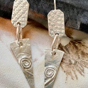 May include: Silver earrings with a hammered rectangular top and a triangular bottom. The bottom features a spiral design. The earrings are connected by a small ring. The earrings are hanging.