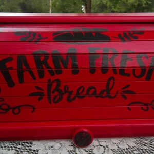 May include: A red wooden bread box with a curved top and a pull-down door. The words "FARM FRESH Bread" are painted in black across the front, with decorative flourishes. The box is designed to store bread and baked goods.