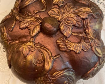 May include: A bronze-colored, decorative pumpkin-shaped container with gold accents. The container features detailed relief carvings of pumpkins, leaves, and vines. The lid has a small, round knob in the center. The overall design is autumnal and decorative.