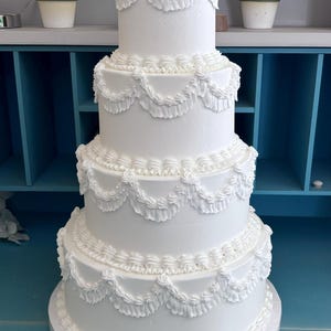 May include: A four-tier white wedding cake decorated with piped frosting and a scalloped design. The cake is on a white cake stand and is in front of a blue shelf.