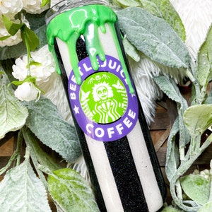 Puede incluir: Un vaso a rayas blancas y negras con un dise&ntilde;o verde goteando en la parte superior. El vaso presenta un logotipo morado y verde que dice "BEETLEJUICE COFFEE". El vaso est&aacute; rodeado de flores blancas y hojas verdes.