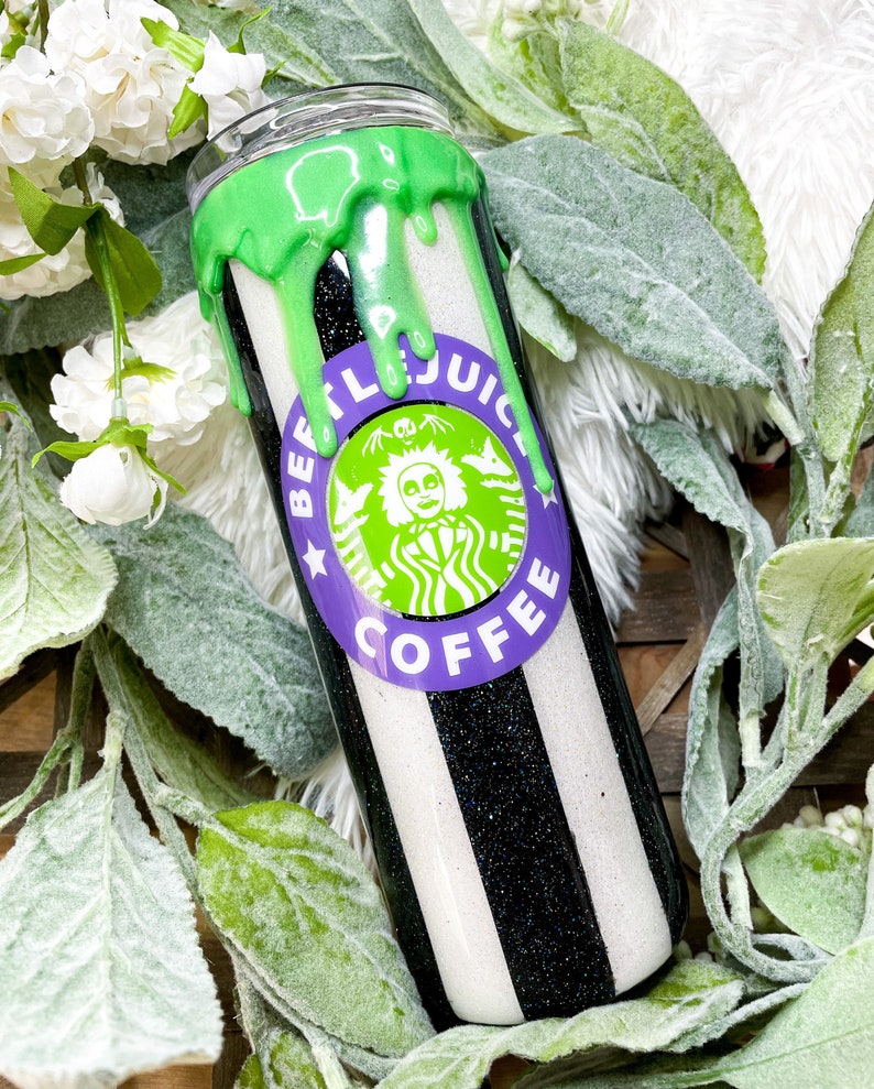 Puede incluir: Un vaso a rayas blancas y negras con un dise&ntilde;o verde goteando en la parte superior. El vaso presenta un logotipo morado y verde que dice "BEETLEJUICE COFFEE". El vaso est&aacute; rodeado de flores blancas y hojas verdes.