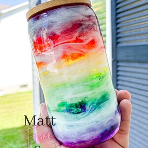 May include: A clear glass tumbler with a rainbow design, featuring red, orange, yellow, green, blue, and purple hues. The tumbler has a bamboo lid and a clear straw. The text "Matt Five Co." is visible on the side.