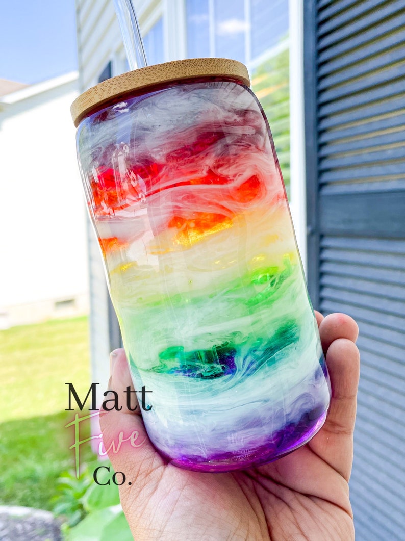 May include: A clear glass tumbler with a rainbow design, featuring red, orange, yellow, green, blue, and purple hues. The tumbler has a bamboo lid and a clear straw. The text "Matt Five Co." is visible on the side.
