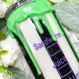 Puede incluir: Un vaso brillante con un dise&ntilde;o de rayas blancas y negras, acentuado con pintura verde ne&oacute;n goteando. El texto "Sandworm Juice Strange & Unusual" est&aacute; impreso en p&uacute;rpura. El vaso tiene una tapa plateada.