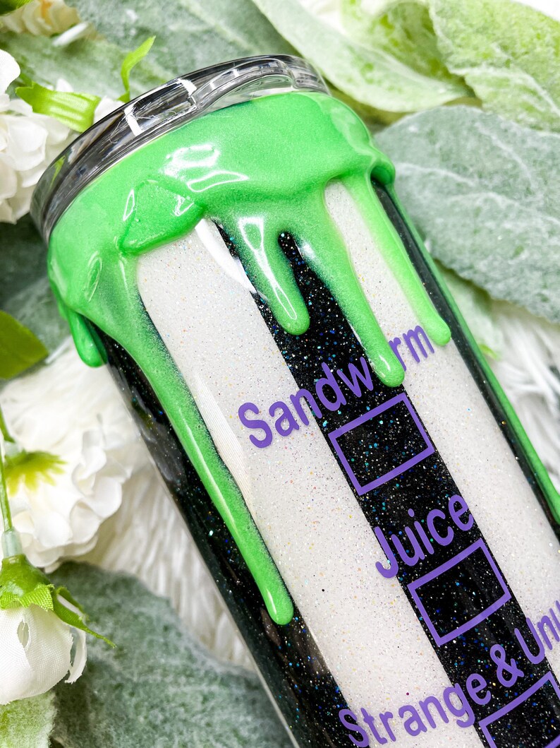 Puede incluir: Un vaso brillante con un dise&ntilde;o de rayas blancas y negras, acentuado con pintura verde ne&oacute;n goteando. El texto "Sandworm Juice Strange & Unusual" est&aacute; impreso en p&uacute;rpura. El vaso tiene una tapa plateada.
