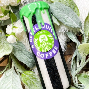 Puede incluir: Un vaso a rayas blancas y negras con un dise&ntilde;o verde goteando en la parte superior. El vaso presenta un logotipo morado y verde que dice "BEETLEJUICE COFFEE" con un personaje de dibujos animados. El vaso est&aacute; rodeado de flores blancas y hojas verdes.