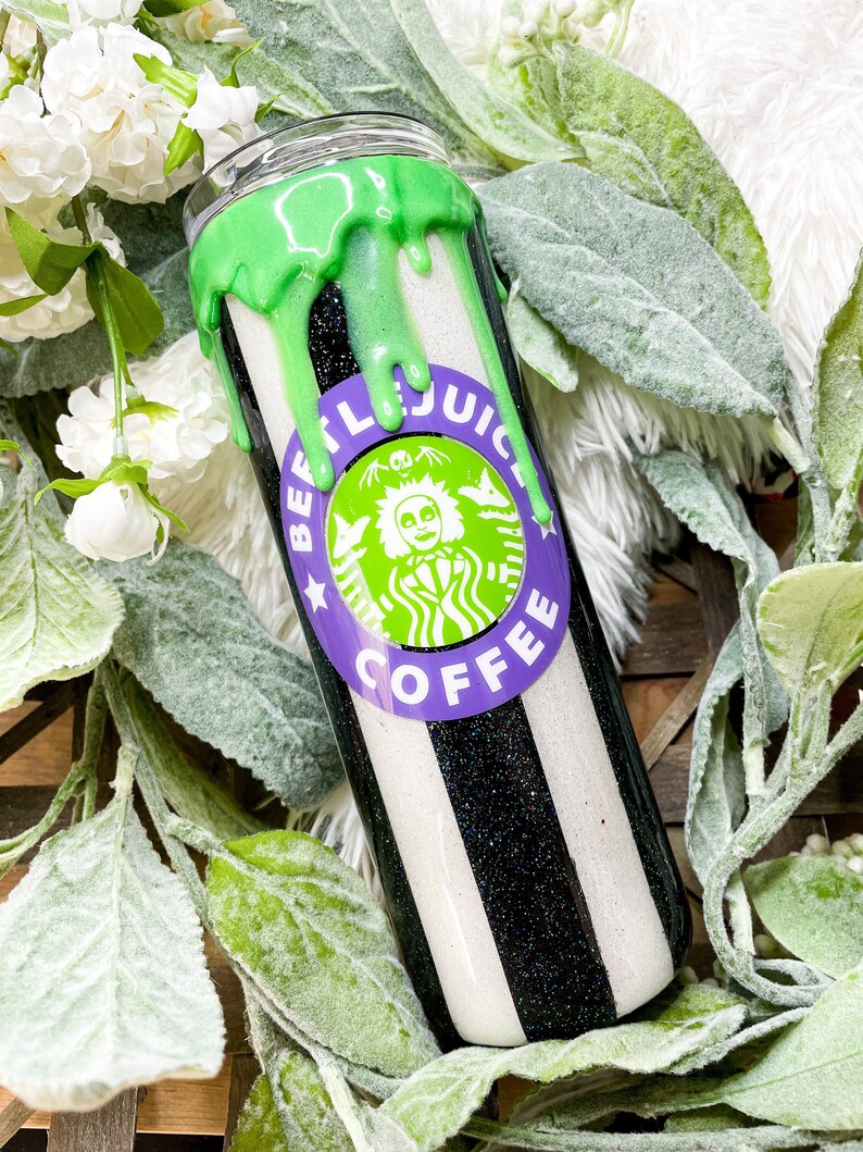 Puede incluir: Un vaso a rayas blancas y negras con un dise&ntilde;o verde goteando en la parte superior. El vaso presenta un logotipo morado y verde que dice "BEETLEJUICE COFFEE" con un personaje de dibujos animados. El vaso est&aacute; rodeado de flores blancas y hojas verdes.
