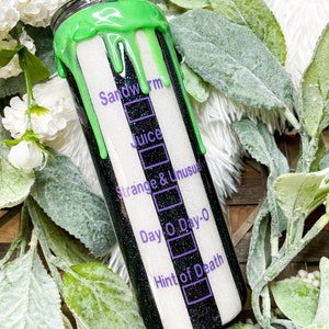 Puede incluir: Un vaso a rayas blancas y negras con un dise&ntilde;o verde goteando en la parte superior. El vaso tiene texto que dice "Sandworm", "Juice", "Strange & Unusual", "Day O Day-0" y "Hint of Death". El vaso est&aacute; rodeado de vegetaci&oacute;n.