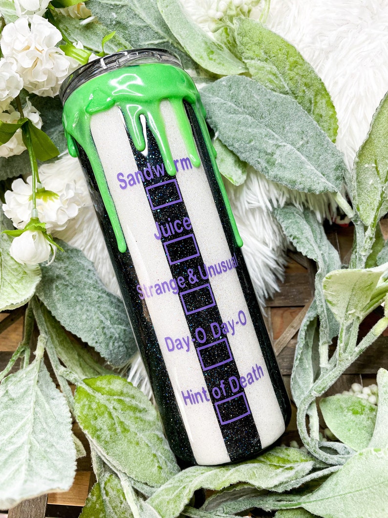 Puede incluir: Un vaso a rayas blancas y negras con un dise&ntilde;o verde goteando en la parte superior. El vaso tiene texto que dice "Sandworm", "Juice", "Strange & Unusual", "Day O Day-0" y "Hint of Death". El vaso est&aacute; rodeado de vegetaci&oacute;n.