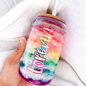 May include: A glass tumbler with a rainbow design and the name "Loren". The tumbler has a bamboo lid and a clear straw. The design features shades of red, orange, yellow, green, blue, and purple. The tumbler is held in a hand.