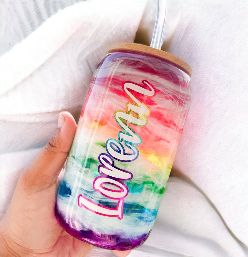 May include: A glass tumbler with a rainbow design and the name "Loren". The tumbler has a bamboo lid and a clear straw. The design features shades of red, orange, yellow, green, blue, and purple. The tumbler is held in a hand.