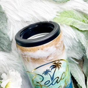 May include: A tumbler with a black rim and a beach-themed design. The tumbler features a gold glitter band, white and blue swirls, and a gold palm tree graphic with the words "Beach Please".