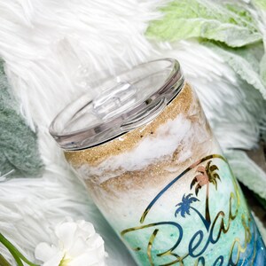May include: A stainless steel tumbler with a clear lid. The tumbler features a beach-themed design with gold glitter, white and tan swirls, and a palm tree graphic with the words "Beach Breeze".