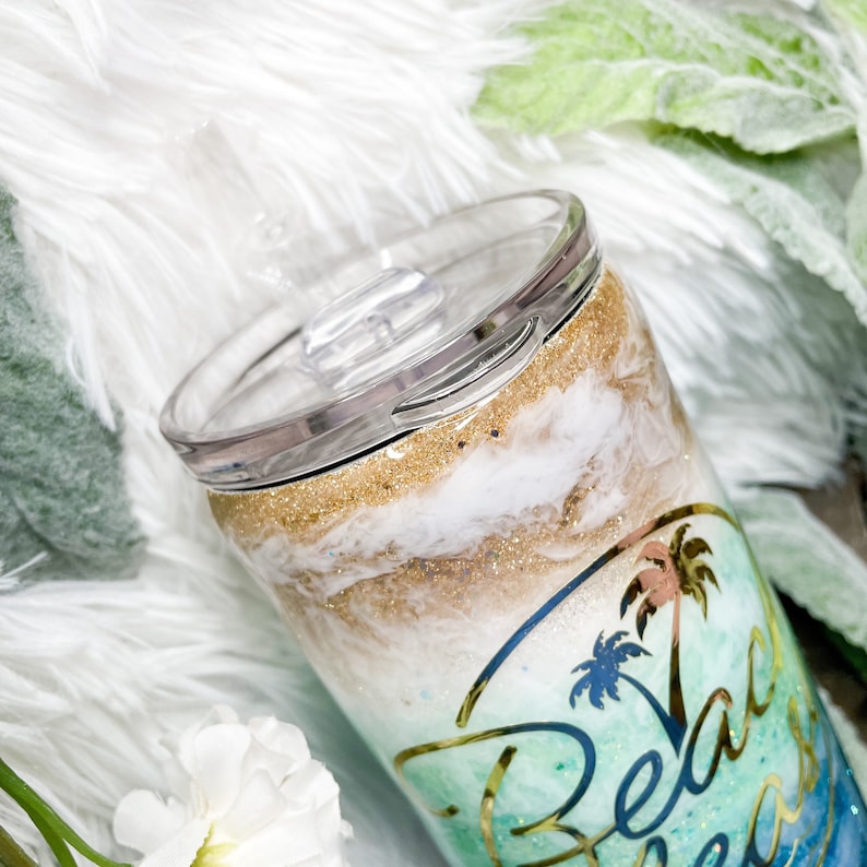 May include: A stainless steel tumbler with a clear lid. The tumbler features a beach-themed design with gold glitter, white and tan swirls, and a palm tree graphic with the words "Beach Breeze".