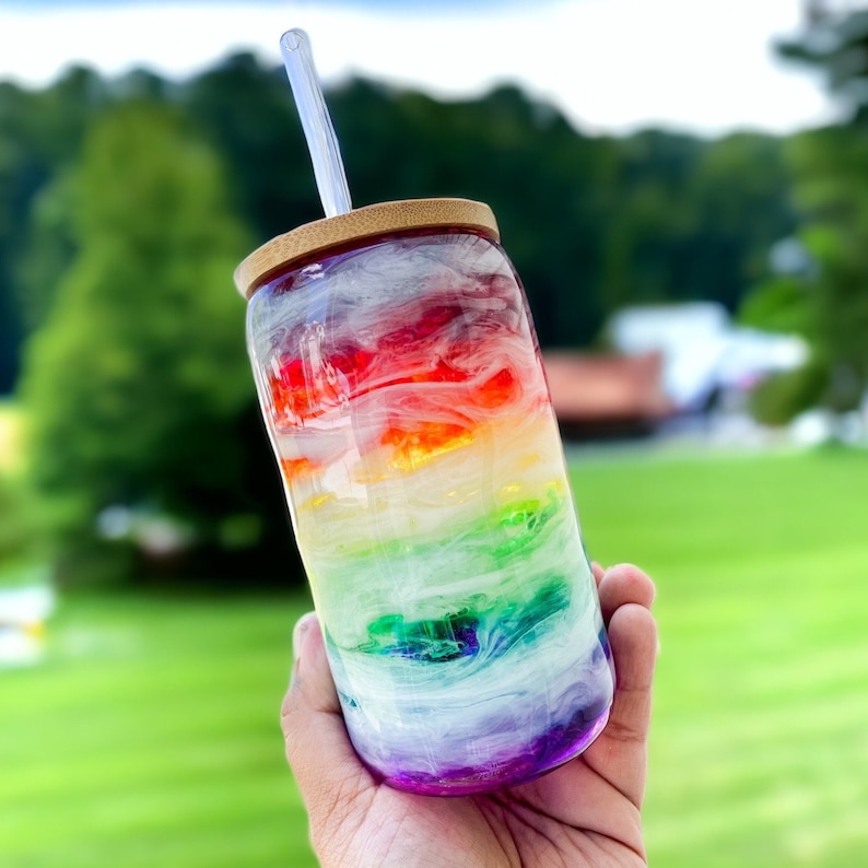 May include: A clear glass tumbler with a rainbow design, a bamboo lid, and a clear straw. The colours blend in a swirling pattern. The tumbler is held against a blurred green background.