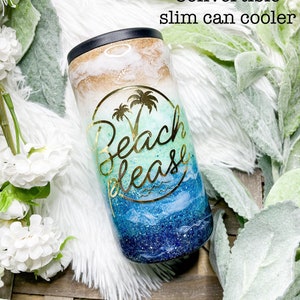 May include: A convertible slim can cooler with a black top and a colorful design. The design features a gold and white top, a turquoise middle with a palm tree graphic, and a blue and navy bottom with the words "Beach Please".
