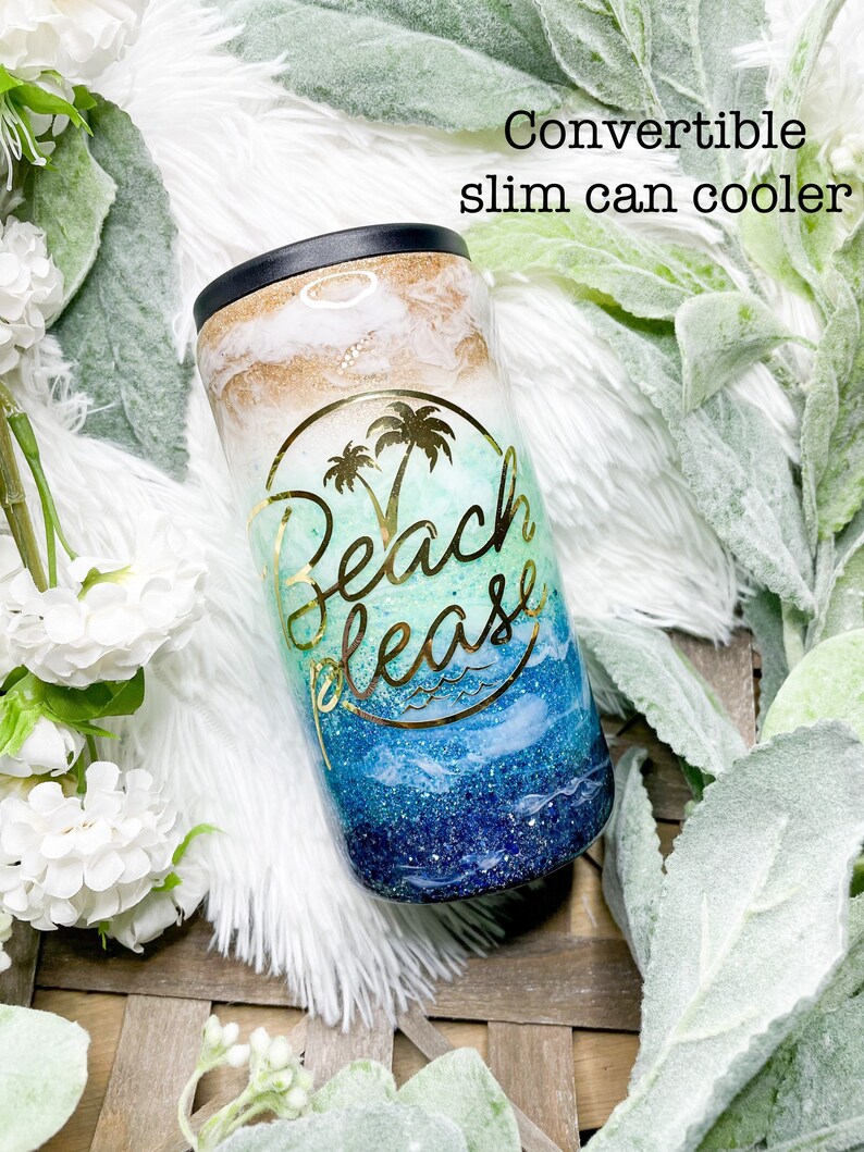 May include: A convertible slim can cooler with a black top and a colorful design. The design features a gold and white top, a turquoise middle with a palm tree graphic, and a blue and navy bottom with the words "Beach Please".