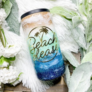 May include: A tumbler with a black rim and a design that says "Beach Please" in gold lettering. The tumbler has a beach-themed design with gold, white, blue, and green colors. The tumbler is cylindrical and appears to be made of metal.