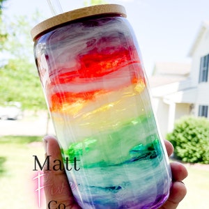 May include: A clear glass tumbler with a rainbow design, a bamboo lid, and a glass straw. The tumbler has a vibrant blend of red, orange, yellow, green, blue, and purple hues. The text "Matt Five Co." is visible on the side.