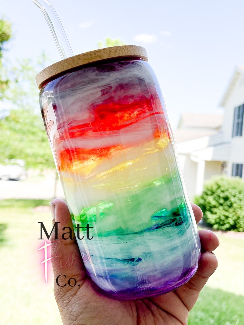May include: A clear glass tumbler with a rainbow design, a bamboo lid, and a glass straw. The tumbler has a vibrant blend of red, orange, yellow, green, blue, and purple hues. The text "Matt Five Co." is visible on the side.