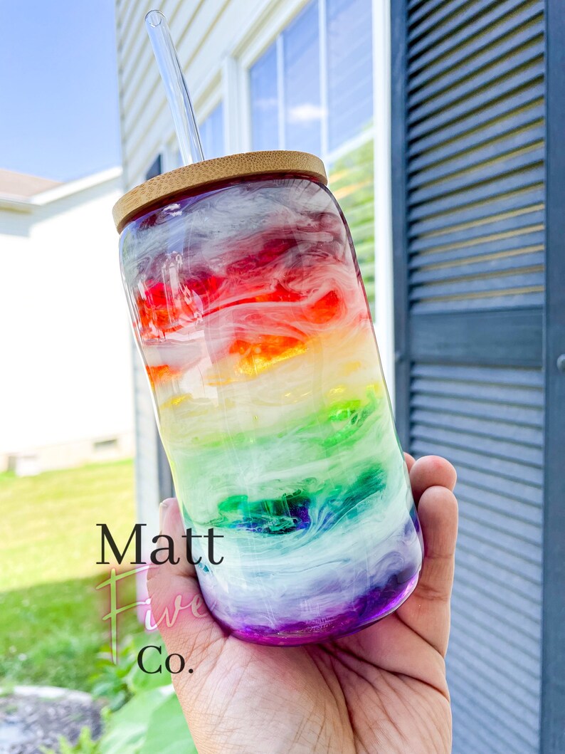 May include: A clear glass tumbler with a rainbow design, a bamboo lid, and a glass straw. The tumbler has a vibrant blend of colours, including red, orange, yellow, green, blue, and purple. The text "Matt Five Co." is printed on the side.