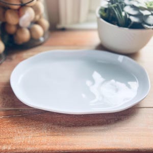 May include: A light blue, oval-shaped ceramic serving platter with an organic, wavy edge. The plate sits on a wooden surface. The background includes a glass jar with brown eggs and a white planter with green plants.