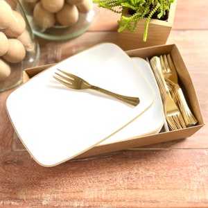 May include: Two white square plates with gold trim, stacked on top of each other. A gold plastic fork is lying on top of the plates. A box of gold plastic forks is in the background.