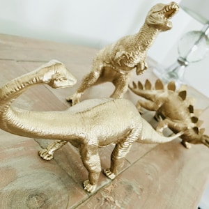 May include: Three gold-painted dinosaur figurines. The dinosaurs are a stegosaurus, a brachiosaurus, and a tyrannosaurus rex. The dinosaurs are arranged on a wooden surface.