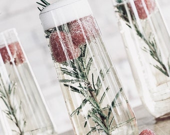 May include: Three clear glass champagne flutes filled with a sparkling beverage, garnished with sprigs of rosemary and red berries. The flutes are arranged on a white surface.