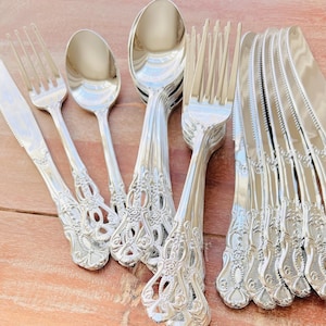 May include: A collection of ornate silver-colored flatware, including forks, spoons, and knives. The handles feature a detailed, decorative design. The flatware is arranged on a wooden surface, showcasing the reflective qualities of the metal.
