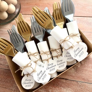 May include: A set of six white napkins wrapped around gold and silver plastic forks, tied with twine and a round tag that says "Merry Christmas 2022".