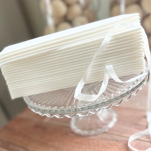 May include: A stack of white fabric face masks tied together with a white ribbon, sitting on a clear glass cake stand.