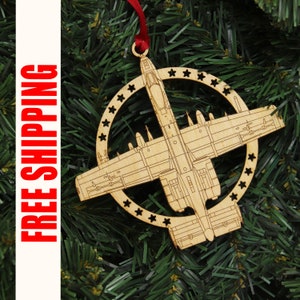 May include: A wooden Christmas ornament shaped like an A-10 Warthog airplane. The ornament is painted in a natural wood color and has a red ribbon attached to the top.
